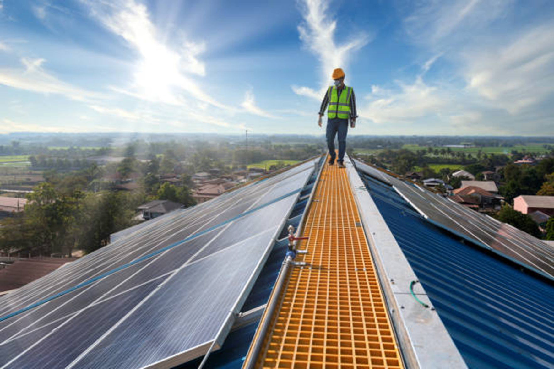 A man in a reflective vest and helmet walking along a rooftop solar panel with the sun shining and some clouds in the sky