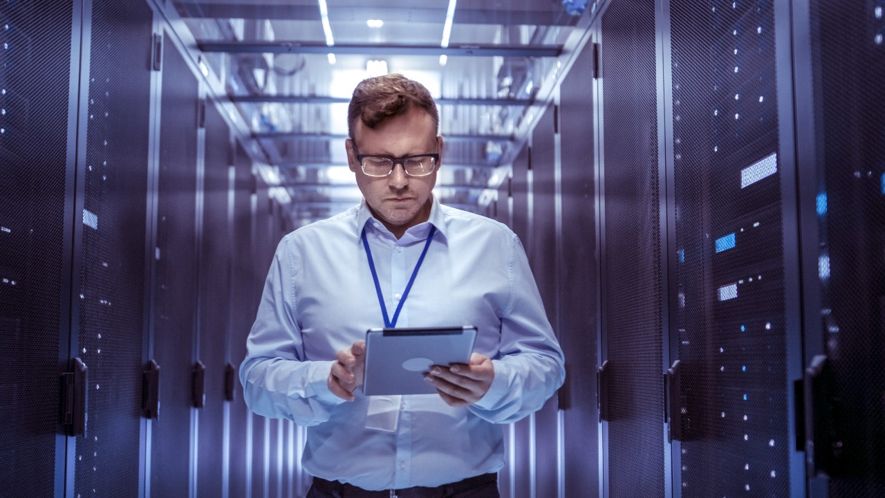 IT Technician Walks Through Rows of Server Racks in Data Center. Simultaneously He Works on a Tablet Computer. IT Technician Walks Through Rows of Server Racks in Data Center. Simultaneously He Works on a Tablet Computer.